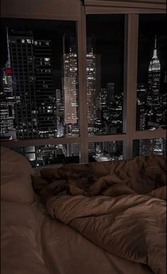 an unmade bed in front of a large window overlooking the city lights at night