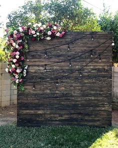 a wooden wall with flowers on it and string lights attached to the side, in front of a fence