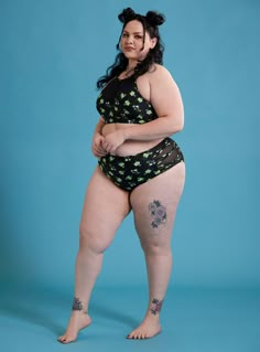 a woman in a black and green swimsuit with tattoos on her legs, standing against a blue background