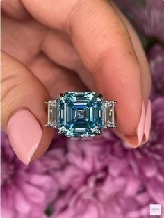 a woman's hand holding an emerald and diamond ring with pink flowers in the background