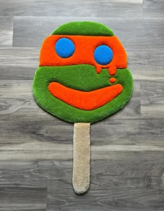 a green, orange and blue lollipop with a face on it sitting on a wooden floor