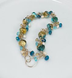 This beautiful bracelet features Czech glass beads, wrapped in 14k Gold Filled wire. The main bracelet consists of Deep Teal Blue beads with a Gold patina and Aurora Borealis luster. I added Deep Ocean Blue, Ivory AB, Aqua, and Clear Gold-Lined beads to create a full bracelet. Everything has been meticulously hand wrapped with 14k Gold Filled wire. The hook & eye clasp is hand forged by me, as always! One of the things I love about cluster bracelets is their beautiful movement. This one is no ex Hand-strung Gold Czech Glass Jewelry, Hand-strung Gold Jewelry With Czech Glass, Gold Czech Glass Jewelry, Hand-strung, Gold Glass Bracelets With Faceted Beads, Gold Hand-strung Czech Glass Bracelets, Gold Czech Glass Hand-strung Bracelet, Gold Bracelets With Faceted Beads, Wire Wrapped Czech Glass Bracelets With Round Beads, Unique Gold Wire Wrapped Beaded Bracelets