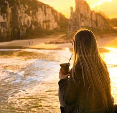 a woman standing on the beach holding a cup in her hand and looking out at the ocean