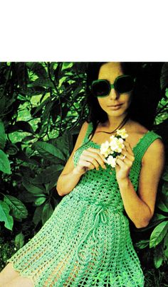 a woman wearing sunglasses and a green crochet dress is holding a white flower