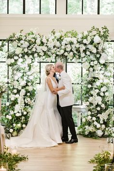 a bride and groom kissing in front of an arch of white flowers at their wedding ceremony