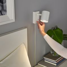 a person is holding a light on the wall next to a bed with books and a potted plant