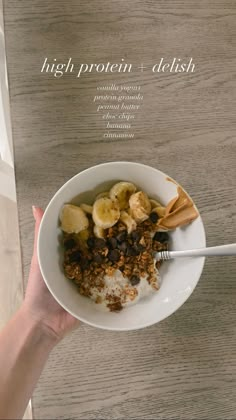 a person holding a bowl of cereal with bananas and granola in it on top of a wooden table