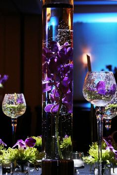 two wine glasses with purple flowers in them on a table next to a tall glass vase