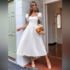 a woman in a white dress standing next to a wall holding a bouquet and posing for the camera