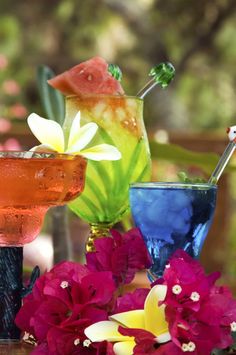 three colorful cocktails sitting on top of a wooden table next to purple and yellow flowers