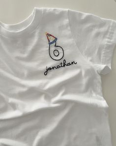 a white t - shirt with the number six on it and a birthday hat embroidered on the front