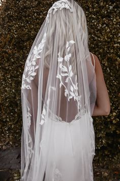 the back of a bride's veil with white flowers on it, in front of a hedge