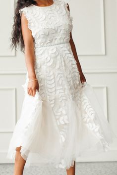 a woman wearing a white dress and heels standing in front of a wall with her hand on her hip