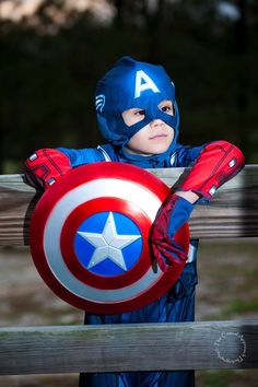 a young boy dressed as captain america holding a shield with both hands on a wooden fence