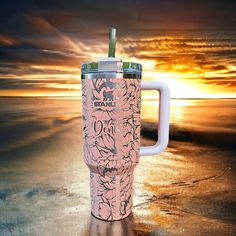 a pink coffee cup sitting on top of a sandy beach next to the ocean at sunset