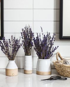 three white vases with lavender flowers in them on a counter top next to scissors