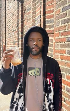 a man in a hoodie is holding up a drink and wearing headphones while standing next to a brick wall