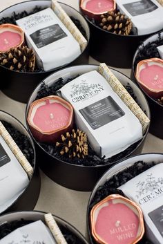 many different types of candles in small black trays on a table with pine cones