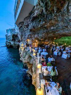 people are sitting at tables next to the ocean in front of a cliff side restaurant