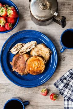 a blue plate topped with french toast and strawberries next to two cups of coffee