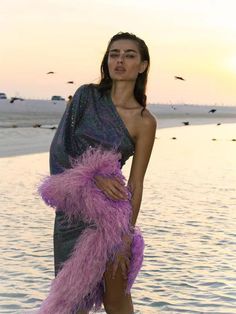 a woman standing in the water wearing a purple feathered dress and matching with sandals