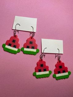 two pairs of red and green earrings with white tags hanging from them on a pink surface