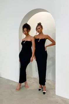 two women standing next to each other in front of a white wall wearing black dresses