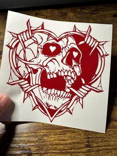 a hand holding up a red and white sticker with a skull in the shape of a heart