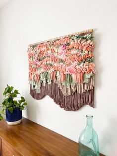 a wall hanging on the side of a wooden shelf next to a vase and plant