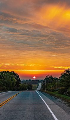 the sun is setting on an empty road with trees in the foreground and clouds in the background