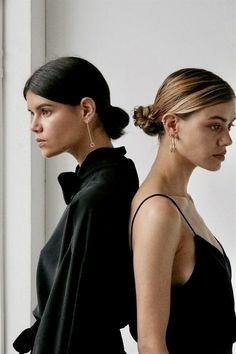 two women standing next to each other in front of a window wearing black dresses and gold earrings