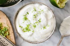 a bowl filled with sour cream next to limes on a cutting board and utensils