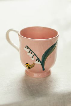 a pink coffee cup sitting on top of a white tablecloth covered table with a green leaf