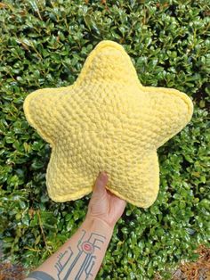 a hand holding up a yellow star shaped pillow in front of some green shrubbery