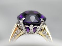 This stunning amethyst cocktail ring has a deep purple amethyst in the center, that really flashes and flickers with vibrant violet reflections! Two rows of square-cut diamonds add sparkle, while round-cut diamonds line the shank of the yellow gold band. Set into a lovely mounting with structurally designed shoulders and a fleur de lis platinum head, this bold statement ring is a beautiful addition to anyone’s collection! Metal: 18K Yellow Gold, Platinum Gem: Amethyst 12.48 Carats Gem Measuremen Exquisite Purple Diamond Rings, Elegant Purple Amethyst Ring With Accent Stones, Luxury Purple Amethyst Ring With Center Stone, Dazzling Purple Amethyst Diamond Ring, Exquisite Purple Jewelry With Center Stone, Purple Cushion Cut Brilliant Ring, Purple Brilliant Cushion Cut Rings, Exquisite Purple Amethyst Ring With Gemstone Accents, Exquisite Diamond Amethyst Ring