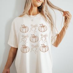 Discover Fall Charm with our Adorable Coquette Core Pumpkins and Bows T-shirt!  Embrace the essence of autumn with the perfect fall tee, a delightful addition to your seasonal wardrobe. Perfectly capturing the spirit of fall, this t-shirt features a charming design of whimsical pumpkins and bows, creating a playful yet elegant look. This shirt offers unparalleled comfort and softness, making it ideal for all-day wear. Its relaxed fit ensures a flattering silhouette that suits any body type. Whet Cute White Tops For Fall, White Short Sleeve Top For Fall, Cute Fall T-shirt With Relaxed Fit, White Relaxed Fit Shirt For Fall, Orange Graphic Tee For Fall, Cute Short Sleeve Tops For Fall, Cute Fall Crew Neck Shirt, Cute White Fall Shirt, Cute Crew Neck Fall Shirt