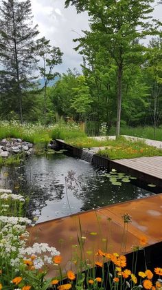 an outdoor pond surrounded by trees and flowers in the foreground, with a wooden walkway leading to it