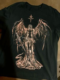 a black shirt with an image of a demon holding a cross on the chest and wings