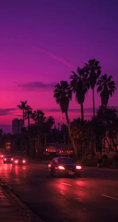 cars driving down the road at night with palm trees and buildings in the back ground