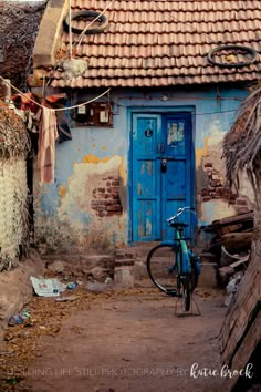 a bicycle parked in front of a blue door on a run down building with thatched roof