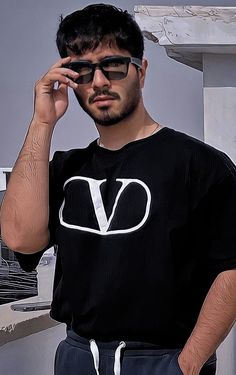 a man in black shirt and sunglasses standing next to a white wall with his hand on his head