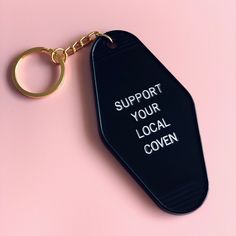 a black keychain with the words support your local coven on it sitting on a pink surface