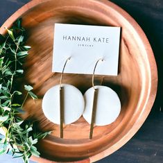 two white round earrings sitting on top of a wooden plate next to a card with the name hannah kate written on it