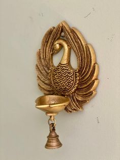 a golden peacock shaped object hanging from the side of a white wall with a bell on it's side