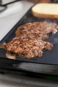 two hamburger patties are being cooked on a griddle with bread in the background