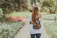 a woman with long red hair walking down a path in the park carrying a backpack