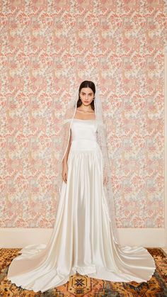 a woman standing in front of a wall wearing a wedding dress and veil with flowers on it