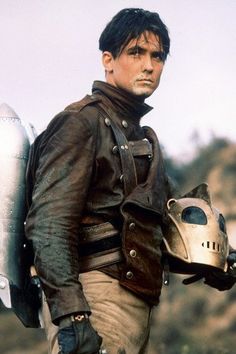 a man in leather jacket holding a motorcycle helmet and looking at the camera while standing next to an airplane