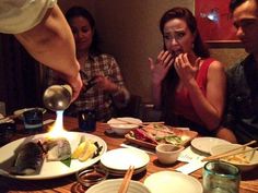 a group of people sitting around a table with plates and bowls on it, one person lighting a candle in the middle