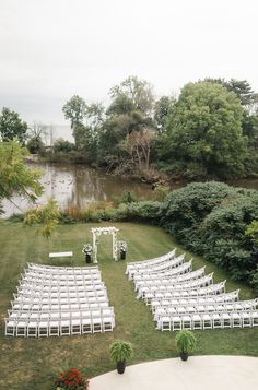 an outdoor ceremony setup with white chairs and flowers on the lawn next to trees, bushes and water